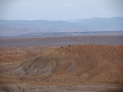 [A dirt hill with rocks over much of it adn two lone pieces of shrubbery at the top. In the distance are canyon walls and mountains.]